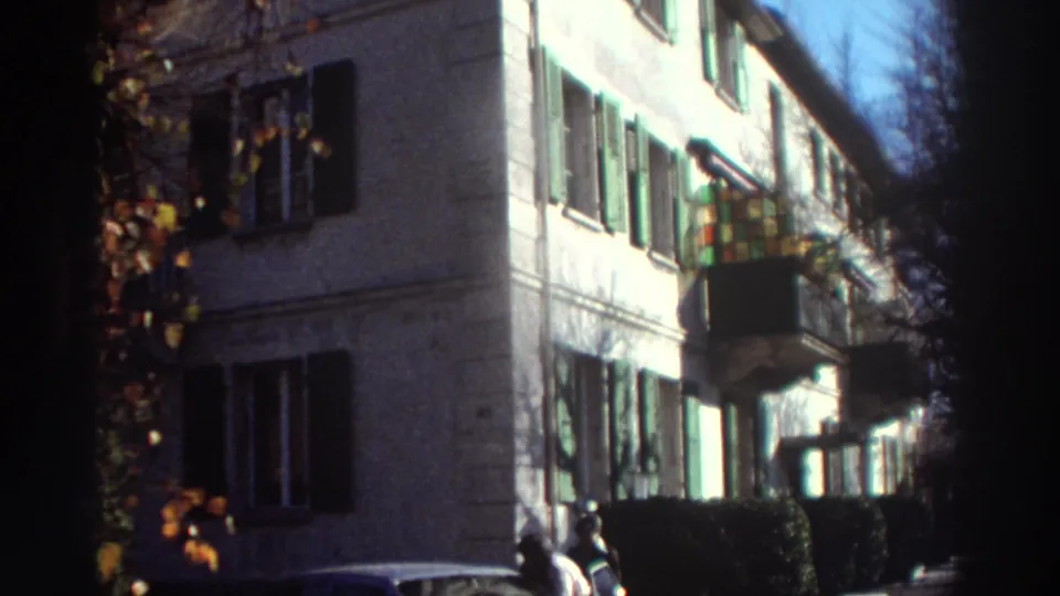 1976 Greece home-movie frame showing a sunlit building facade with green shutters, a small balcony, shrubs, and two people near the street.