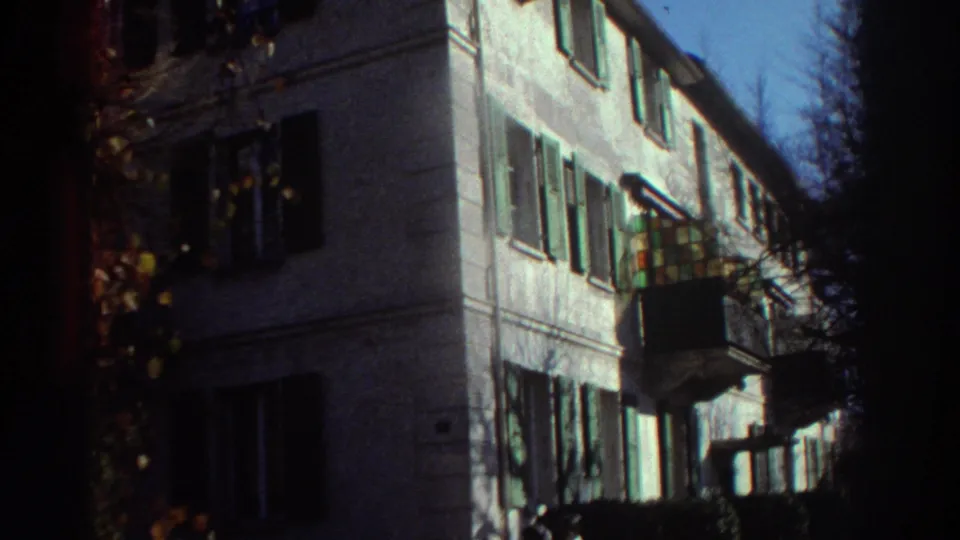 1976 Greece 8mm frame with a closer view of the shuttered residential building, leafy edge, balcony, and a figure partly visible near the bottom.