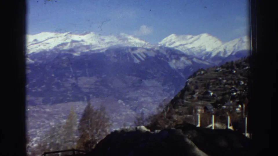 1976 Greece home-movie frame with snow-covered mountains, a valley, bare winter trees, and a dark hillside structure or slope.
