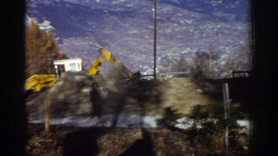 1976 Greece 8mm frame showing a yellow excavator near a small white structure, with a valley backdrop and a blurred person in the foreground.