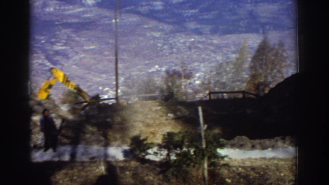 Yellow excavator working near a snowy mountain road with an adult figure in the foreground.