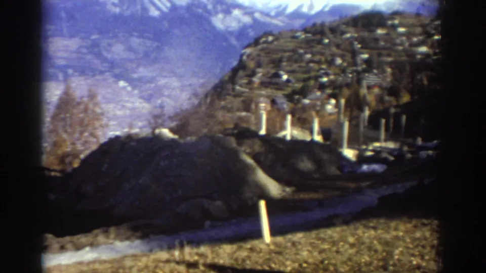 1976 Greece frame with hillside buildings, distant winter mountains, bare trees, roadside posts, and a dark mound in the foreground.