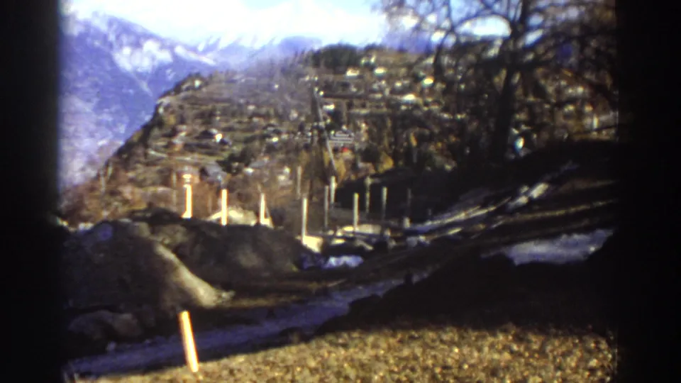 1976 Greece home-movie frame showing a hillside settlement, snow-capped mountains, bare branches, roadside posts, and rough winter terrain.