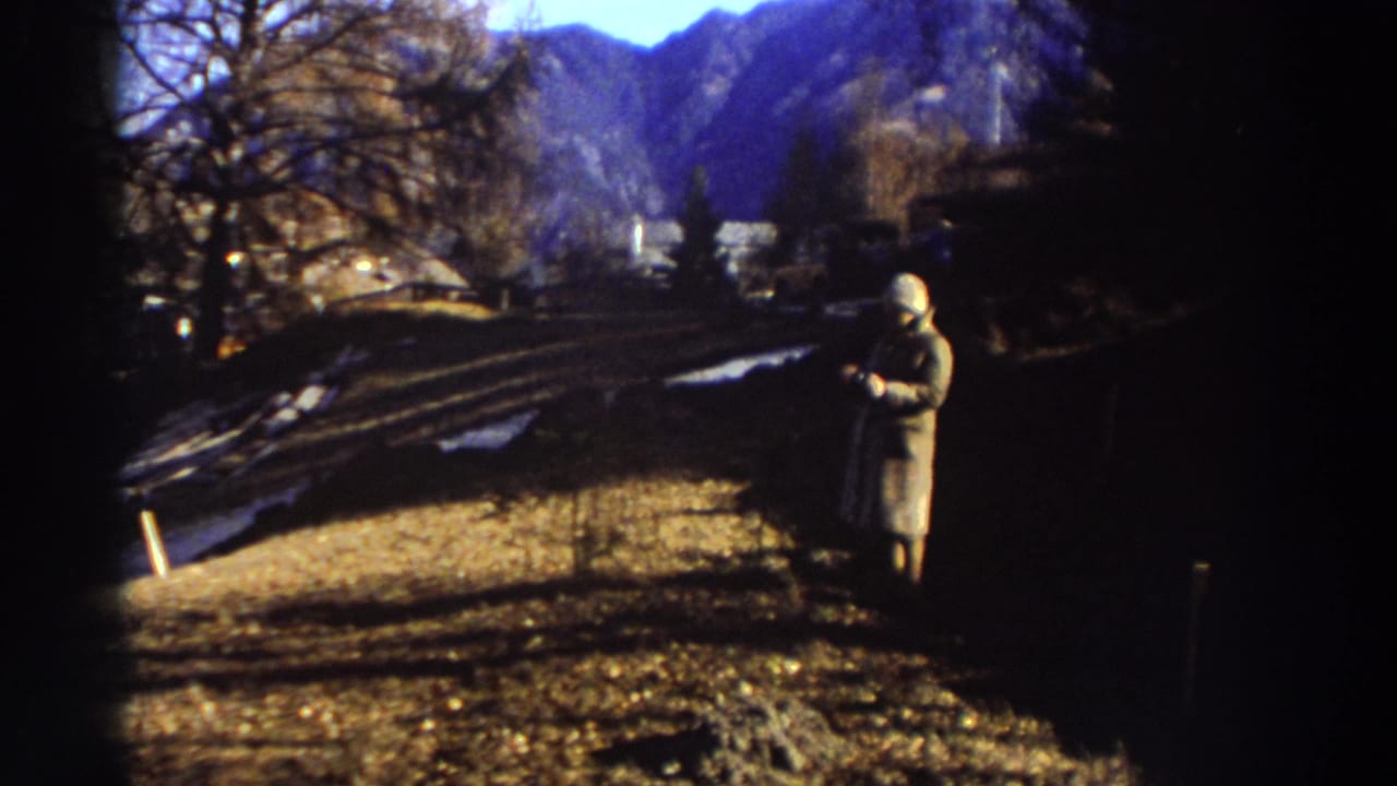 1976 Greece home-movie frame showing a trench-coated person standing on a dry hillside with leafless trees and mountains in the distance.