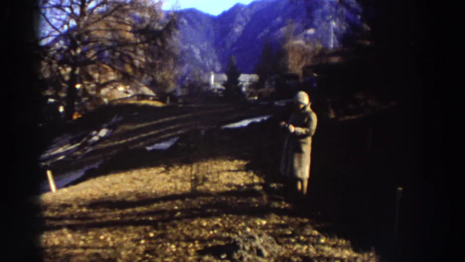 1976 Greece home-movie frame showing a trench-coated person standing on a dry hillside with leafless trees and mountains in the distance.
