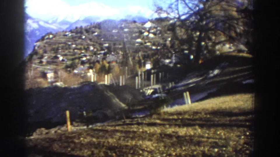 The camera looks over a hillside toward a Greek mountain village, with snow patches, trees, and distant slopes visible in soft 8mm color.