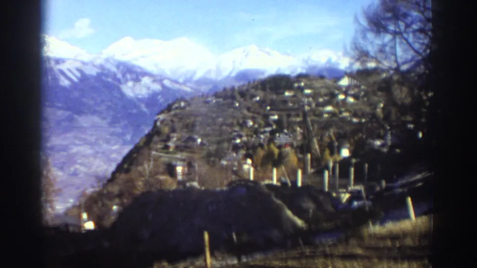 Wide 1976 Greece home-movie view of a mountain village on a slope, with a valley and snow-capped peaks under a bright sky.