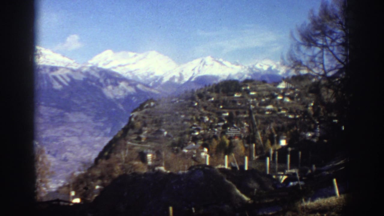 1976 Greece 8mm frame showing a hillside village below snow-capped mountains, with bare trees and dark film edges framing the view.