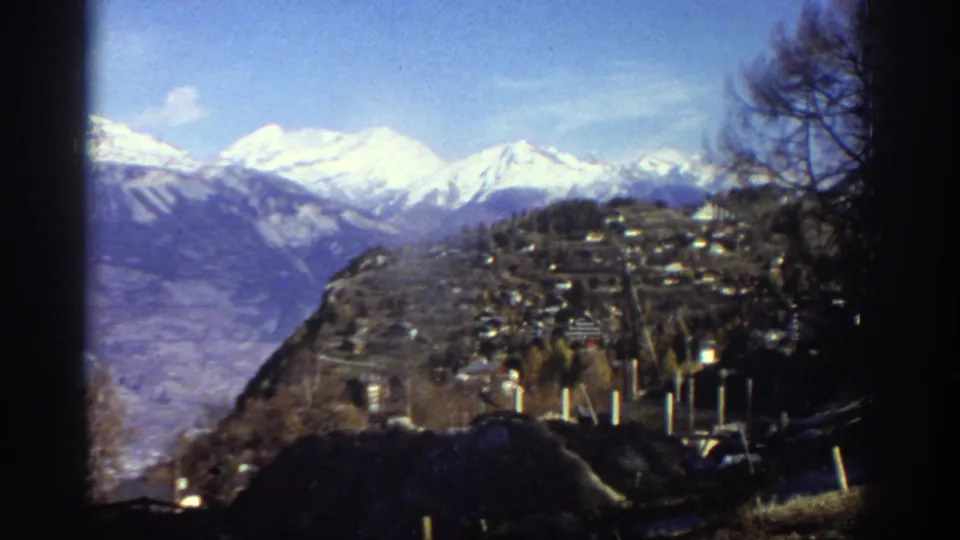 1976 Greece 8mm frame showing a hillside village below snow-capped mountains, with bare trees and dark film edges framing the view.