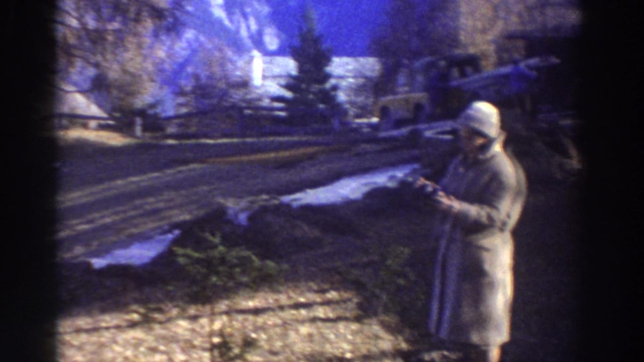 1976 Greece home-movie frame of a woman in winter clothing standing by a snowy hillside road, appearing to hold a small camera.