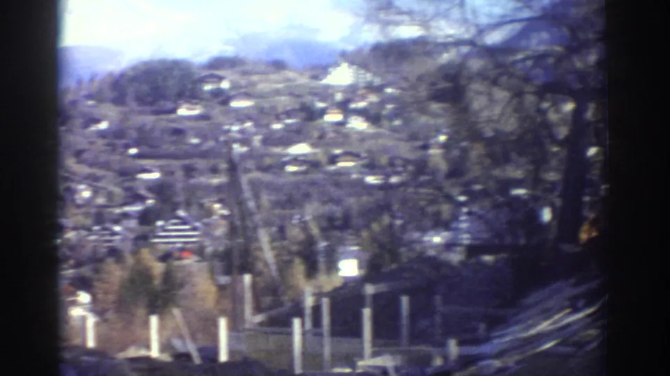 1976 Greece home-movie frame looking across a hillside village, with white fence posts, bare trees, and distant mountain slopes visible.