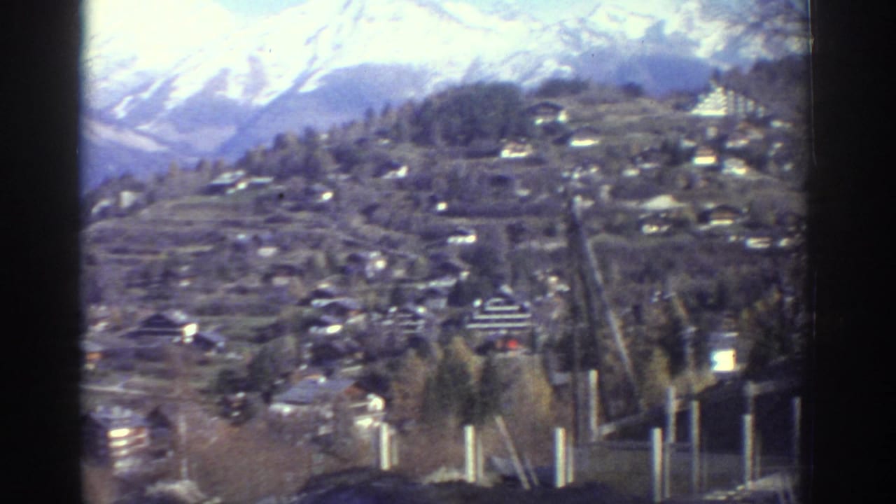 1976 Greece 8mm view of a hillside settlement with terraced slopes and snowy mountains in the distance.