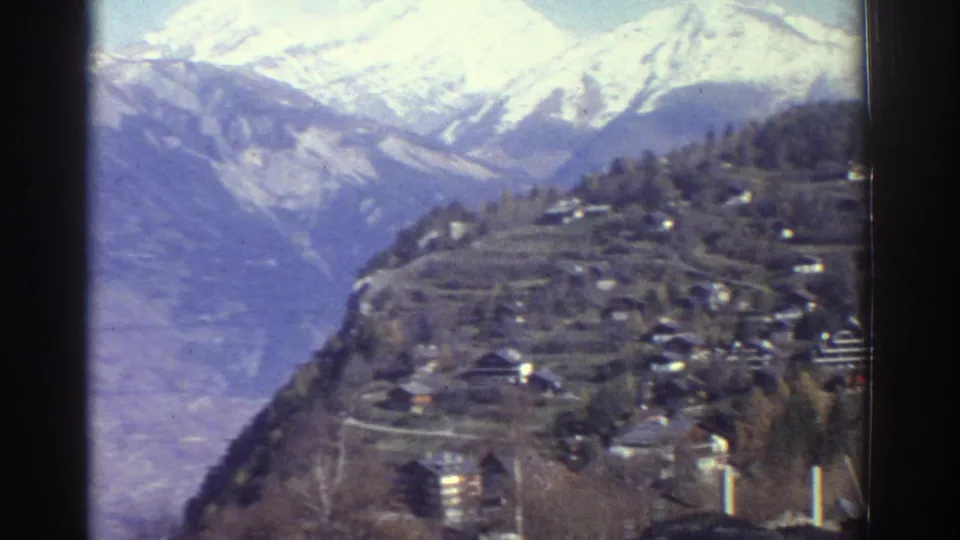 1976 Greece home-movie frame showing a steep mountain valley, hillside houses, trees, and distant snow-covered peaks.