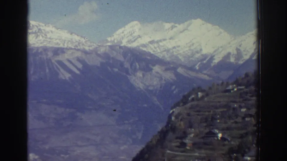 1976 Greece 8mm frame of snow-capped mountains behind a shadowed hillside and distant rural buildings.
