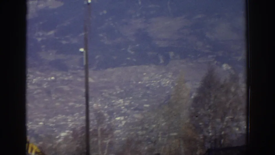 1976 Greece home-movie frame showing snow-capped mountains, a valley settlement, bare trees, and a utility pole through a window.