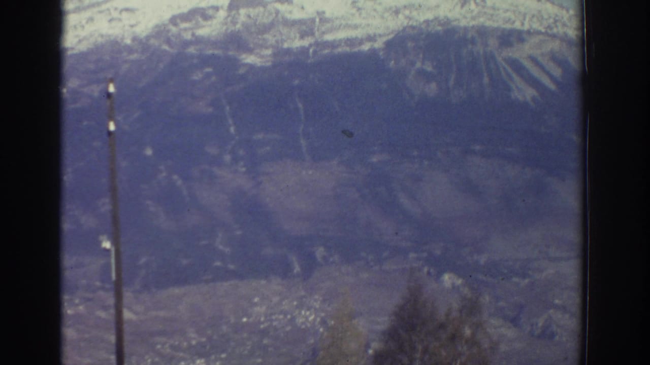 1976 Greece landscape frame with snowy mountain ridges, a valley below, bare foreground branches, and a utility pole.