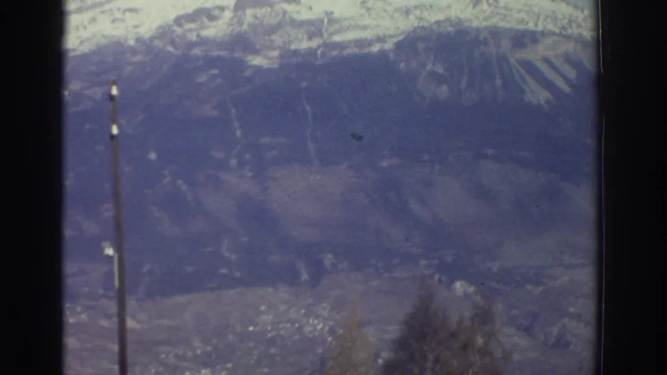 1976 Greece landscape frame with snowy mountain ridges, a valley below, bare foreground branches, and a utility pole.
