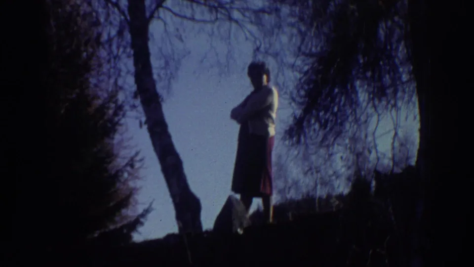 1976 Greece home-movie frame showing a woman in a light sweater and dark skirt standing with arms crossed near trees.