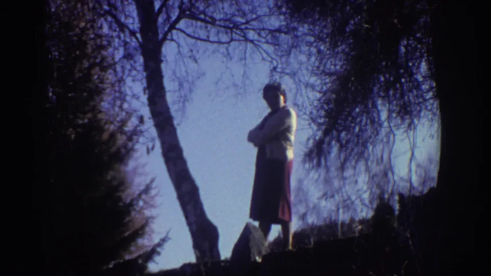 1976 Greece home-movie frame of a woman outdoors near a leaning tree, wearing a light top and dark skirt against a clear sky.