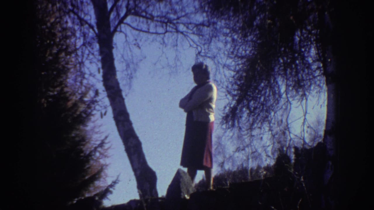 In 1976 Greece home-movie footage, an elderly woman in a light sweater and dark skirt stands on a wooded rocky rise.