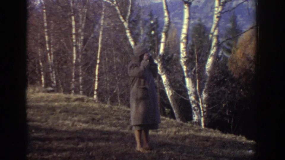 In the Greece 1976 footage, an elderly woman in a coat and hat stands on a grassy hillside with a camera.