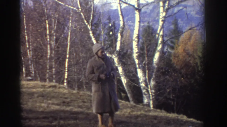 A coated elderly woman holds a camera while standing on a wooded hillside in 1976 Greece home-movie footage.