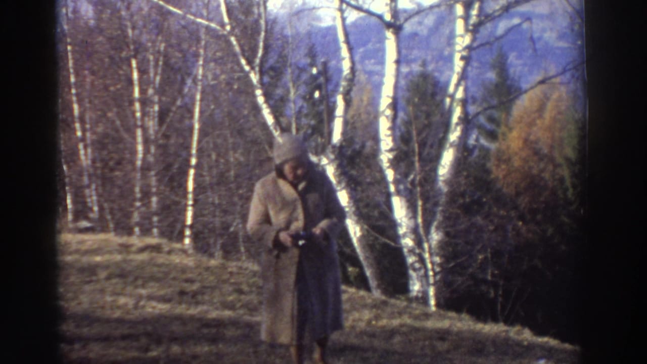 1976 Greece 8mm frame of a woman in a long coat and hat walking on a grassy slope with birch-like trees and mountains behind her.