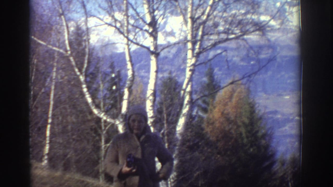 1976 Greece home-movie frame showing a cloaked woman holding a camera among trees, with blue-toned mountains in the distance.