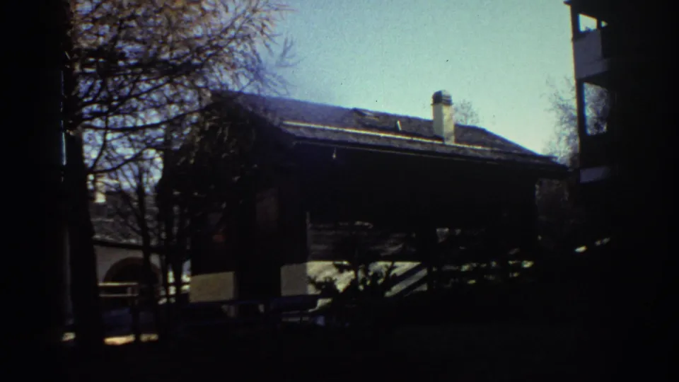 1976 Greece home-movie frame showing an exterior wooden building or chalet partly shaded by trees, with a mountain backdrop.