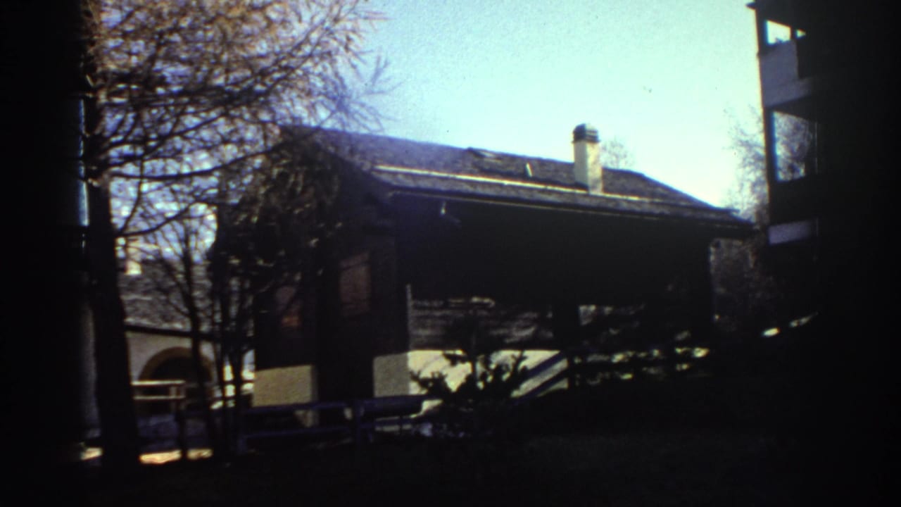 1976 Greece home-movie frame showing a rustic house, bare trees, and strong shadowed edges in a mountain village setting.