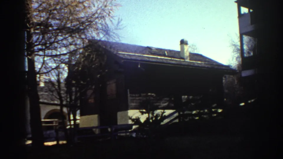 1976 Greece home-movie frame showing a rustic house, bare trees, and strong shadowed edges in a mountain village setting.
