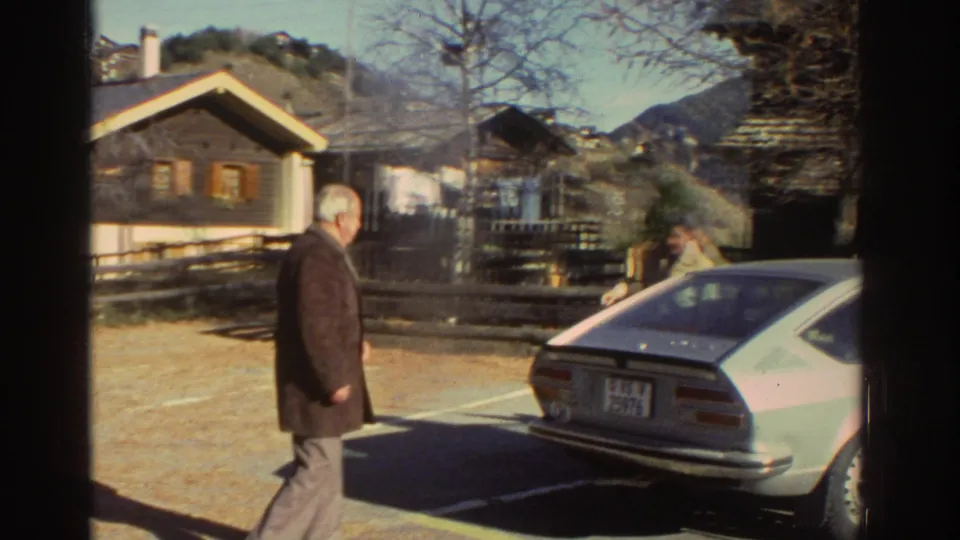 An adult man walks near a parked silver hatchback, rustic houses, fencing, and mountains in 1976 Greece home-movie footage.