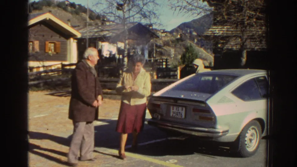 Two adults stand beside a parked silver hatchback in a sunlit Greek village scene from 1976 home-movie footage.