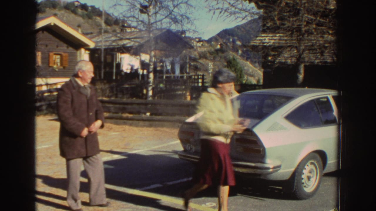 1976 Greece home-movie frame showing an older man and woman near a parked light-colored coupe with wooden houses and mountains behind them.
