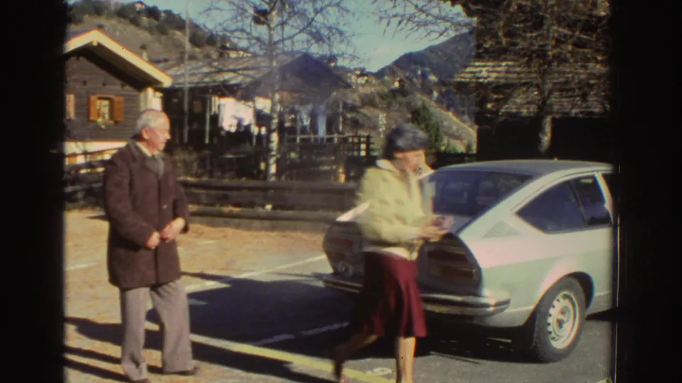 1976 Greece home-movie frame showing an older man and woman near a parked light-colored coupe with wooden houses and mountains behind them.