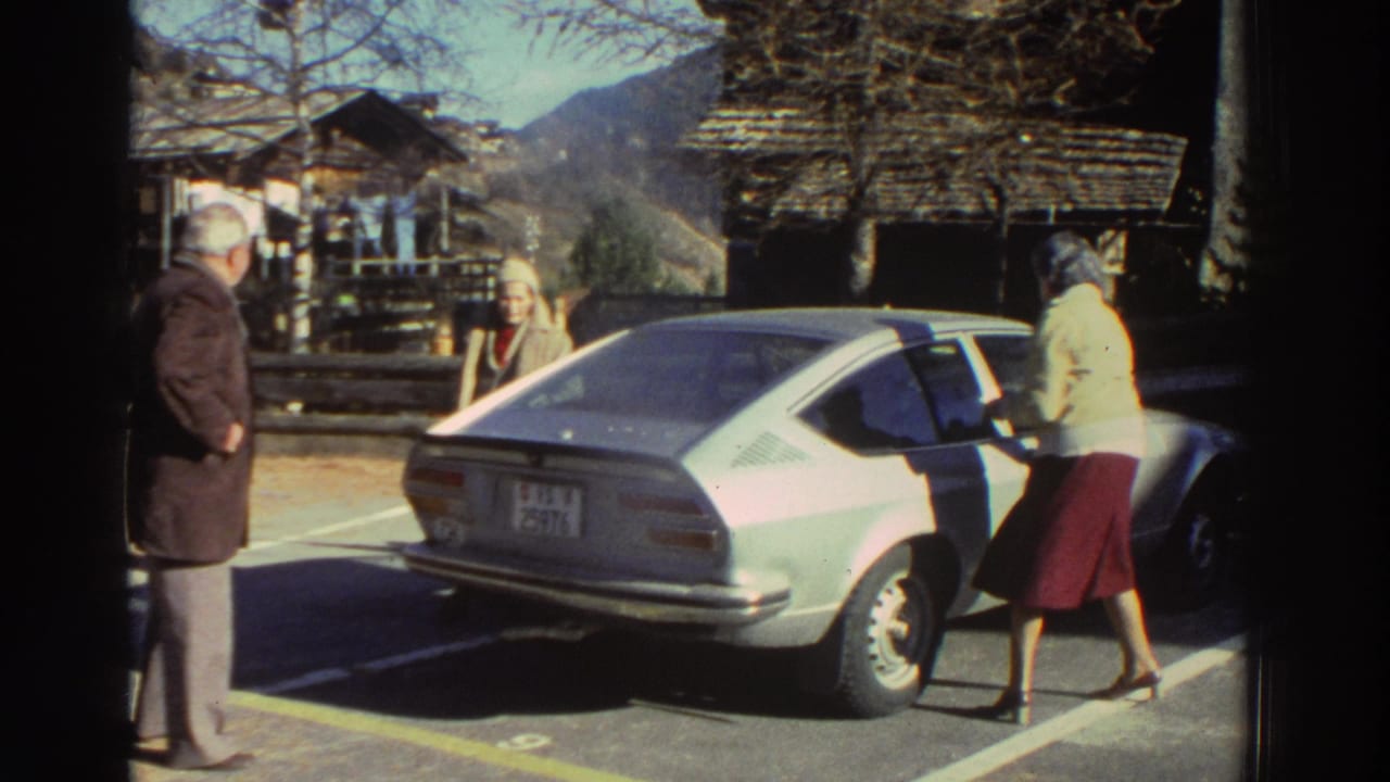 Older adults stand around the rear of a light-colored coupe in a mountain village parking area.