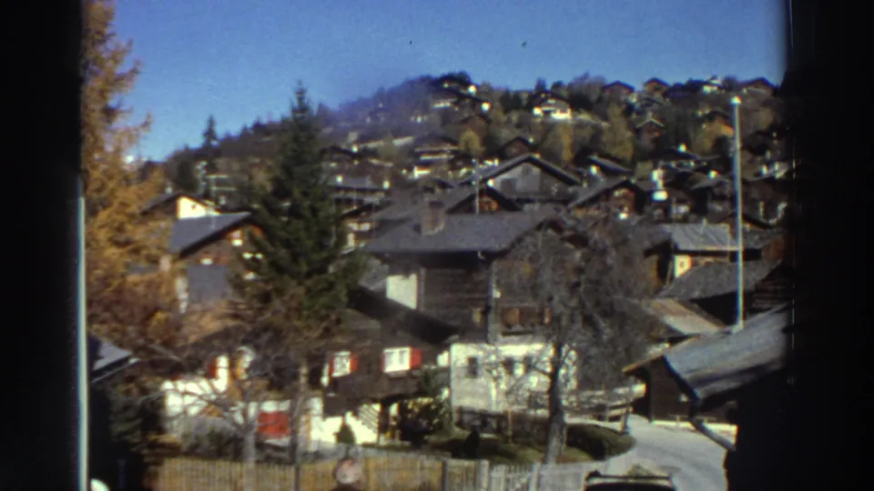 1976 Greece frame showing a hilly residential village with wooden houses, bare trees, a road, and mountains in the background.