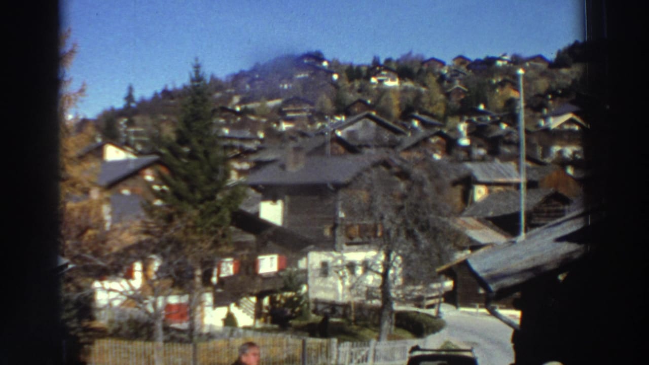 Hillside village homes with trees, fences, and a narrow road under a blue sky.