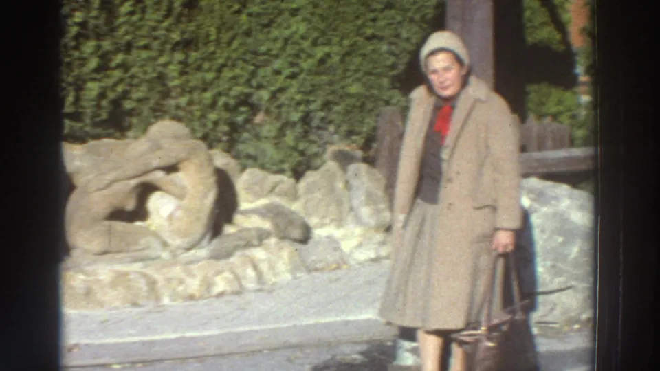 1976 Greece home-movie frame of an adult woman in a beige coat and hat posing near a stone sculpture and hedge.
