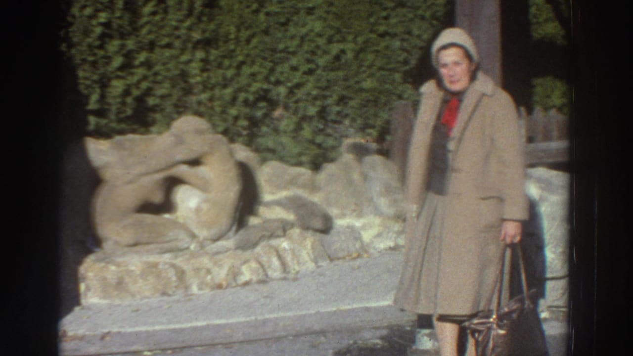 In 1976 Greece home-movie footage, a woman in a beige coat and knit hat stands beside an outdoor stone sculpture.