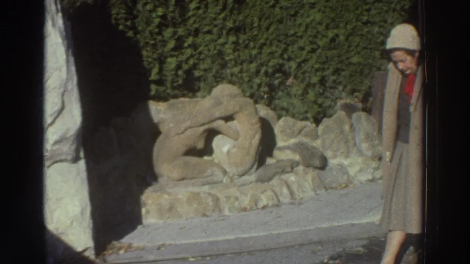 The woman appears at the right edge of the 1976 Greece frame, looking down beside the stone sculpture and hedge.