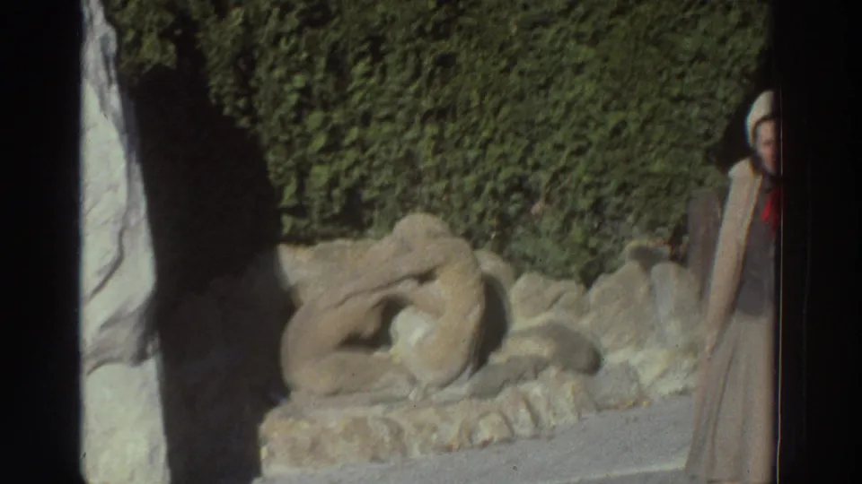 The camera frames the stone sculpture and hedge while the woman in a beige coat is partly visible at the right edge.