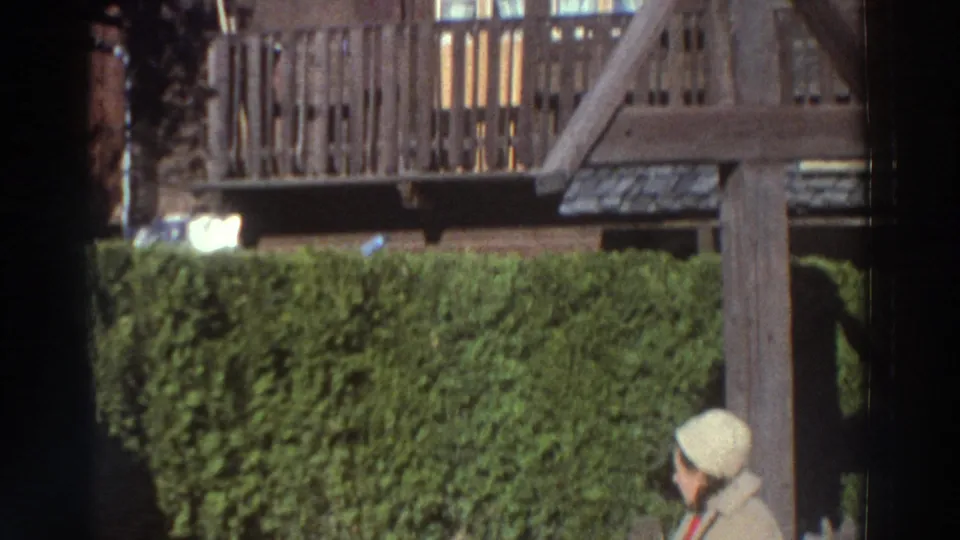 The woman turns near the hedge in 1976 Greece footage, with a wooden structure visible in the background.