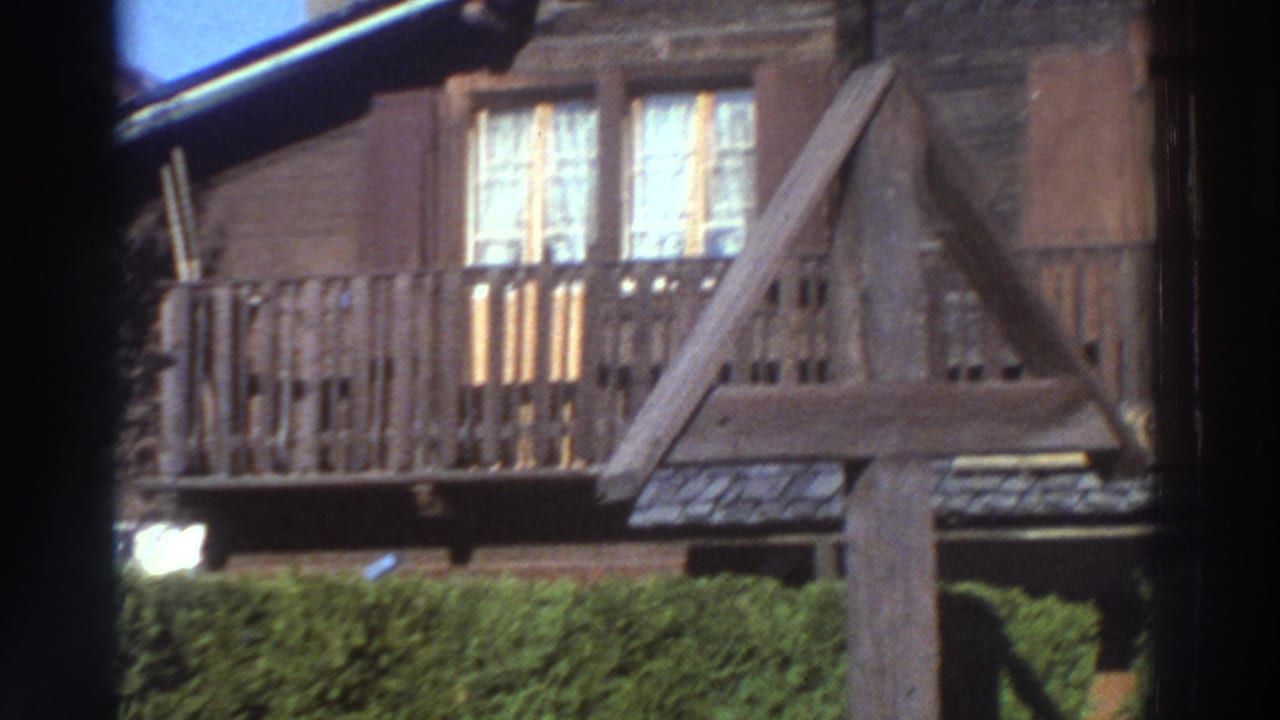 1976 Greece 8mm frame showing a brown timber house exterior with balcony rails, shuttered windows and lace curtains seen through a dark edge.