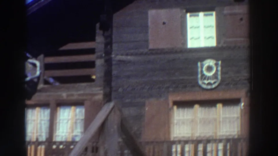 1976 Greece 8mm frame of the wooden facade, with a bright curtained window, closed shutters, balcony rail and a circular emblem on the wall.