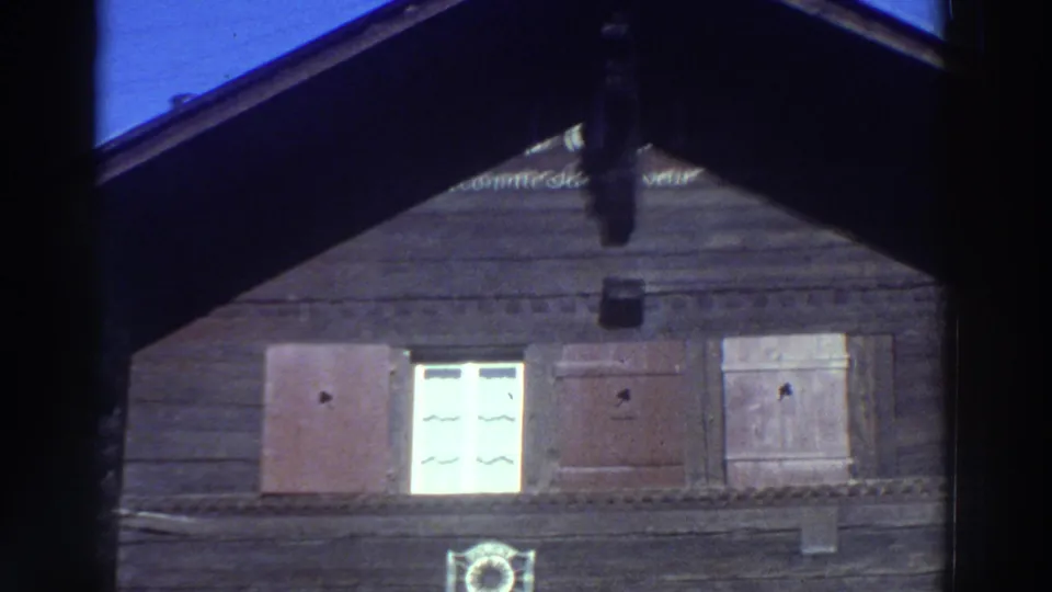 1976 Greece 8mm frame showing the upper story of a brown wooden building with dark roof edge, shutters, a lit window and a circular emblem.