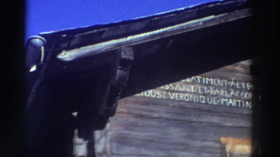 1976 Greece 8mm frame with a dark roof edge crossing the view and partially readable painted lettering on the wooden facade below.