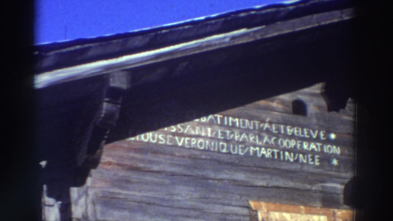 1976 Greece home-movie frame showing a weathered wooden facade with French text, dark roof eaves, and blue sky above.