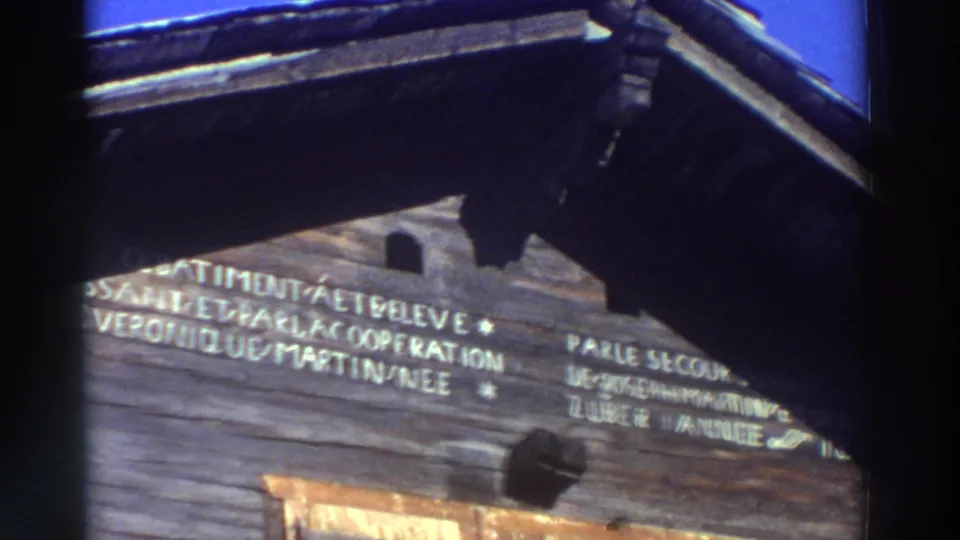 1976 Greece home-movie frame of a rustic wooden building, with French words painted or carved across the upper facade.
