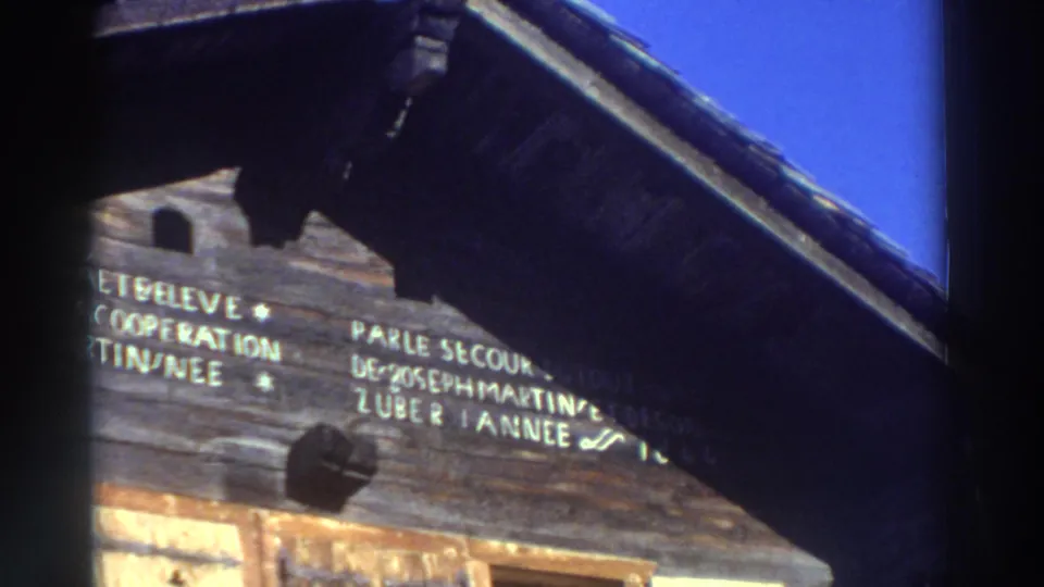1976 Greece home-movie frame showing an angled view of weathered planks, partial French lettering, and a dark sloping roof.